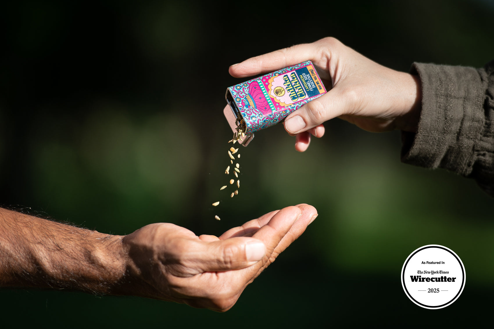 Person pouring a fennel seed mix from a colorful metal tin into another person's hand against a blurred natural background